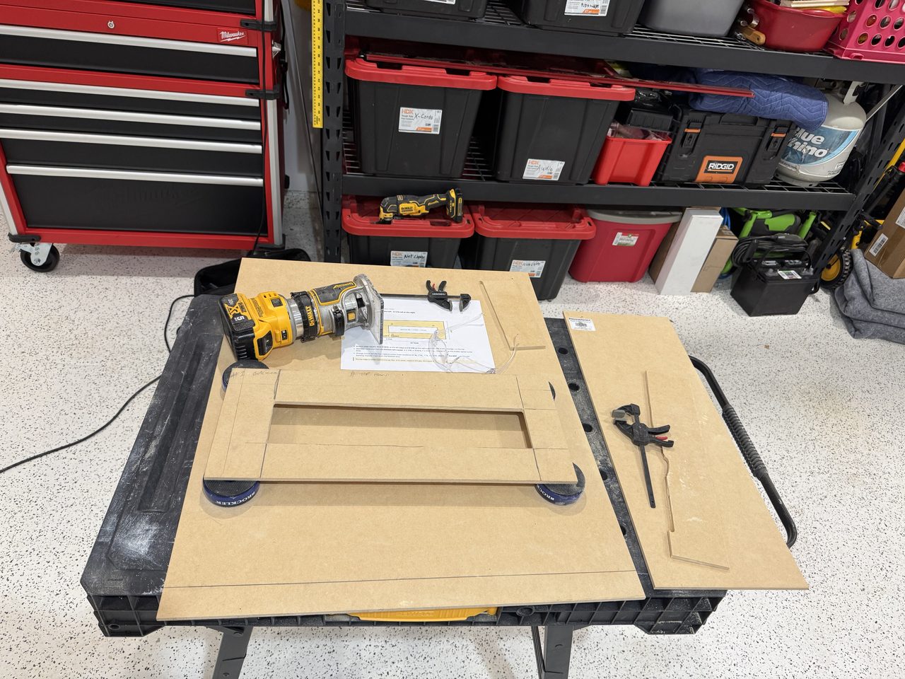 A folding plastic worktable in a garage, covered with sheets of pale brown MDF. In the middle a rectangular panel with a long rectangular hole cut through it rests on blue bench pads. Beside it are a yellow compact router, a printed drawing, safety glasses and two small clamps; to the right is a narrow offcut with a clamp on it. Behind are storage shelves of red-lidded bins, with an oscillating multi-tool on one shelf, and a red tool chest.