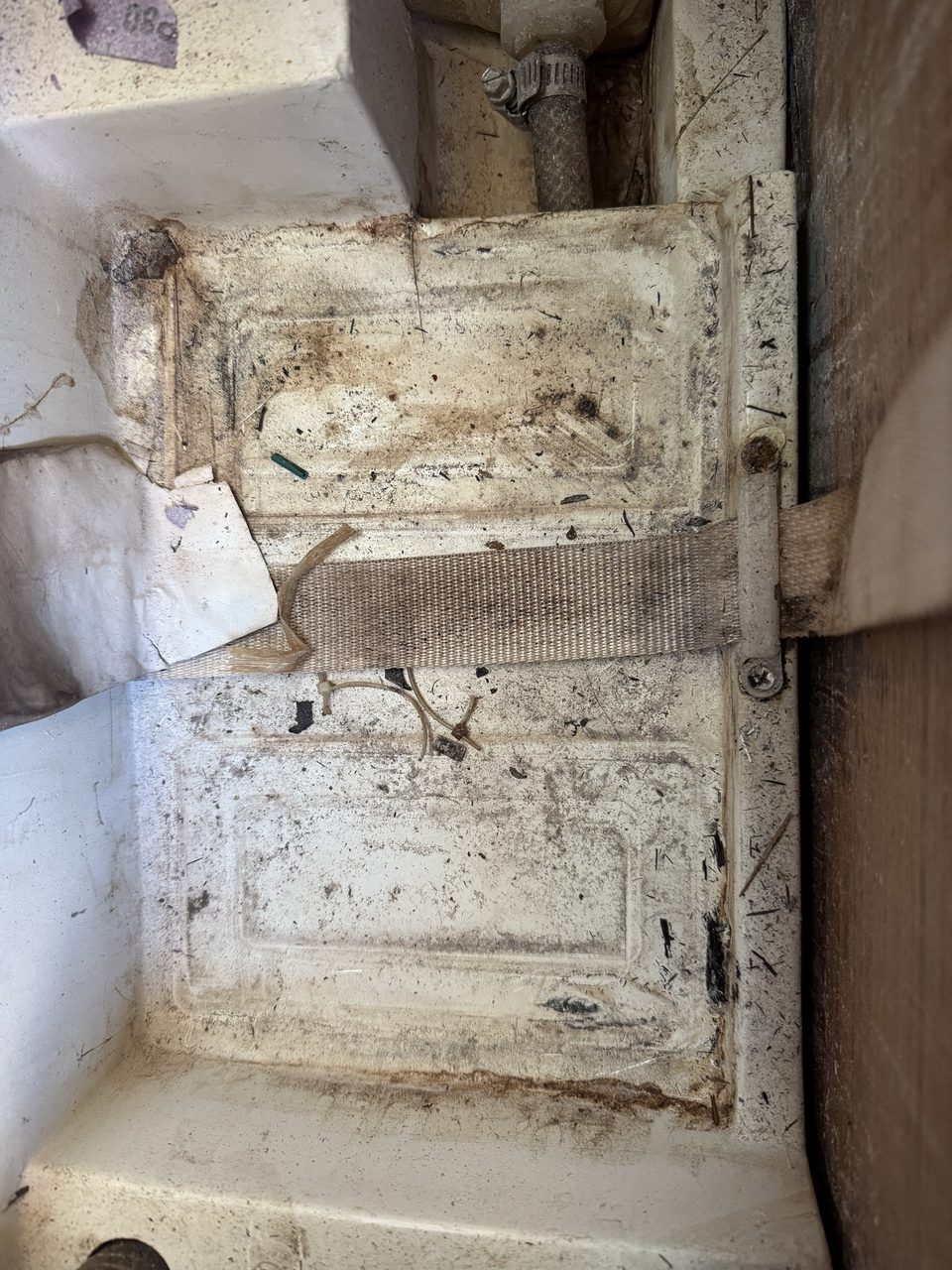 A filthy moulded battery well in a boat, coated in dirt and debris, with a frayed and dirty webbing strap across it
