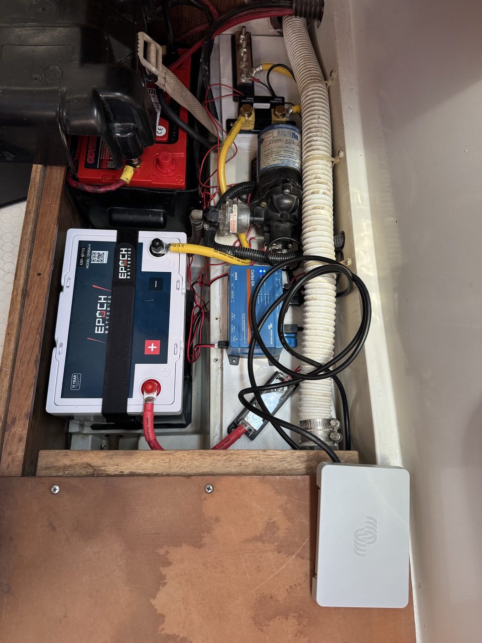 Looking down into a boat battery compartment with a white lithium battery, an older battery, a pump, a blue control unit, cabling in red and yellow, and a small display panel resting on a wooden lid
