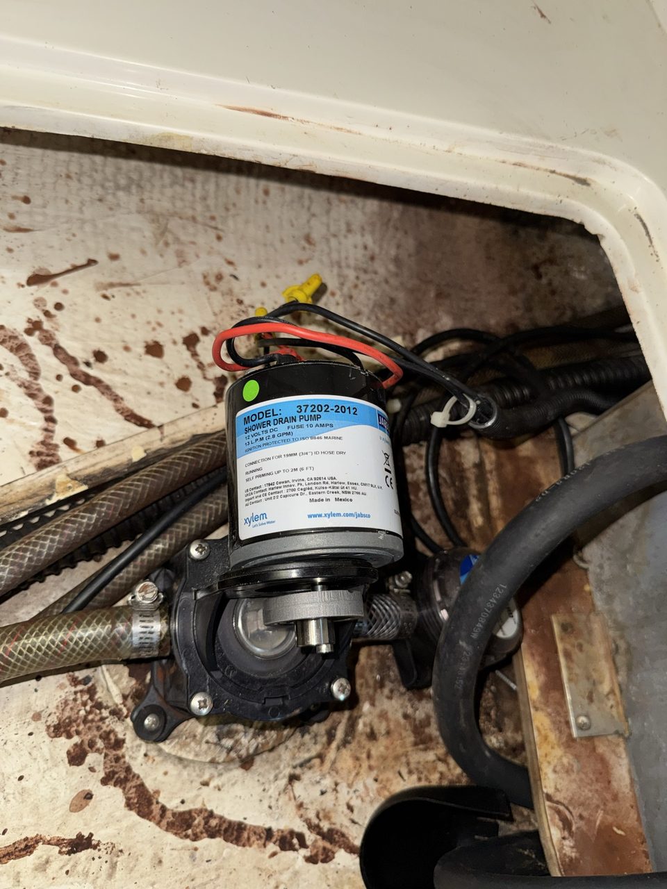 A small black and white electric diaphragm pump mounted on a stained white fibreglass surface inside a boat locker, with red and black wires and a clear pump housing