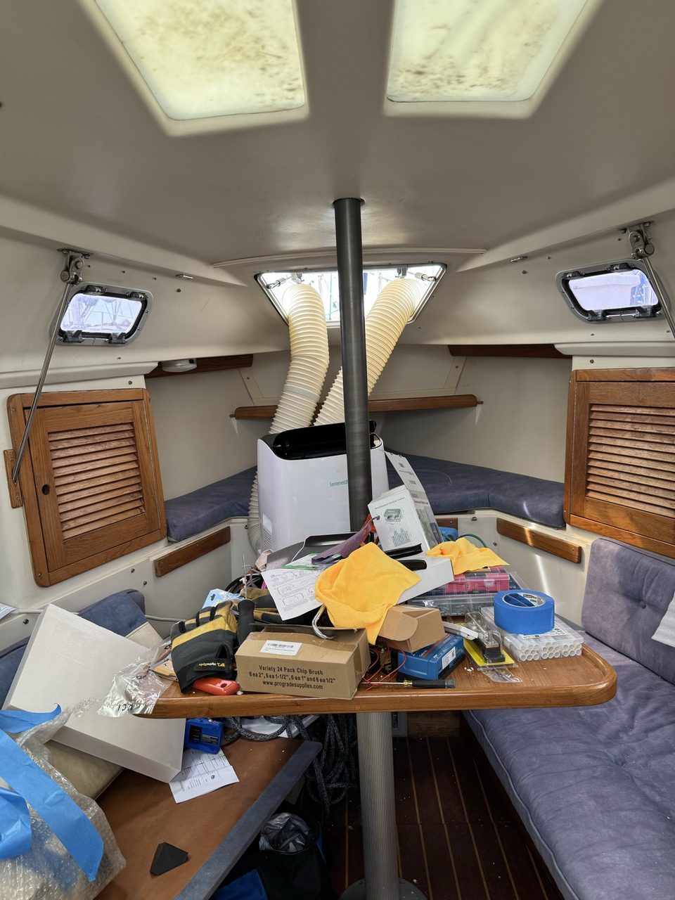 The interior of a sailboat looking forward, with a portable air conditioner on the forward berth and two ducts rising through an open deck hatch, and the saloon table covered in tools and packaging