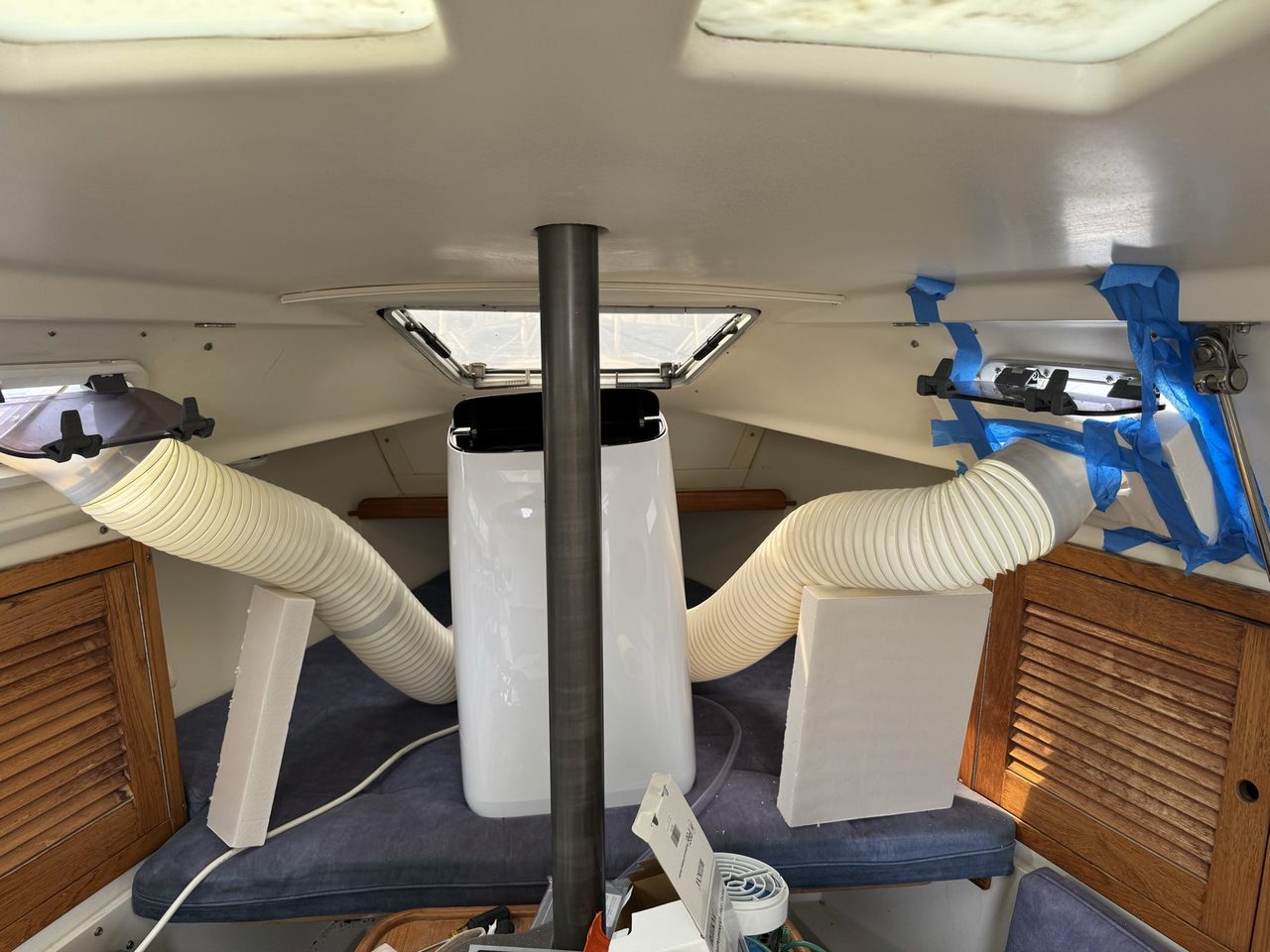 Looking forward inside a sailboat cabin at a portable air conditioner between two settees, with a duct running from each side up to an open port light