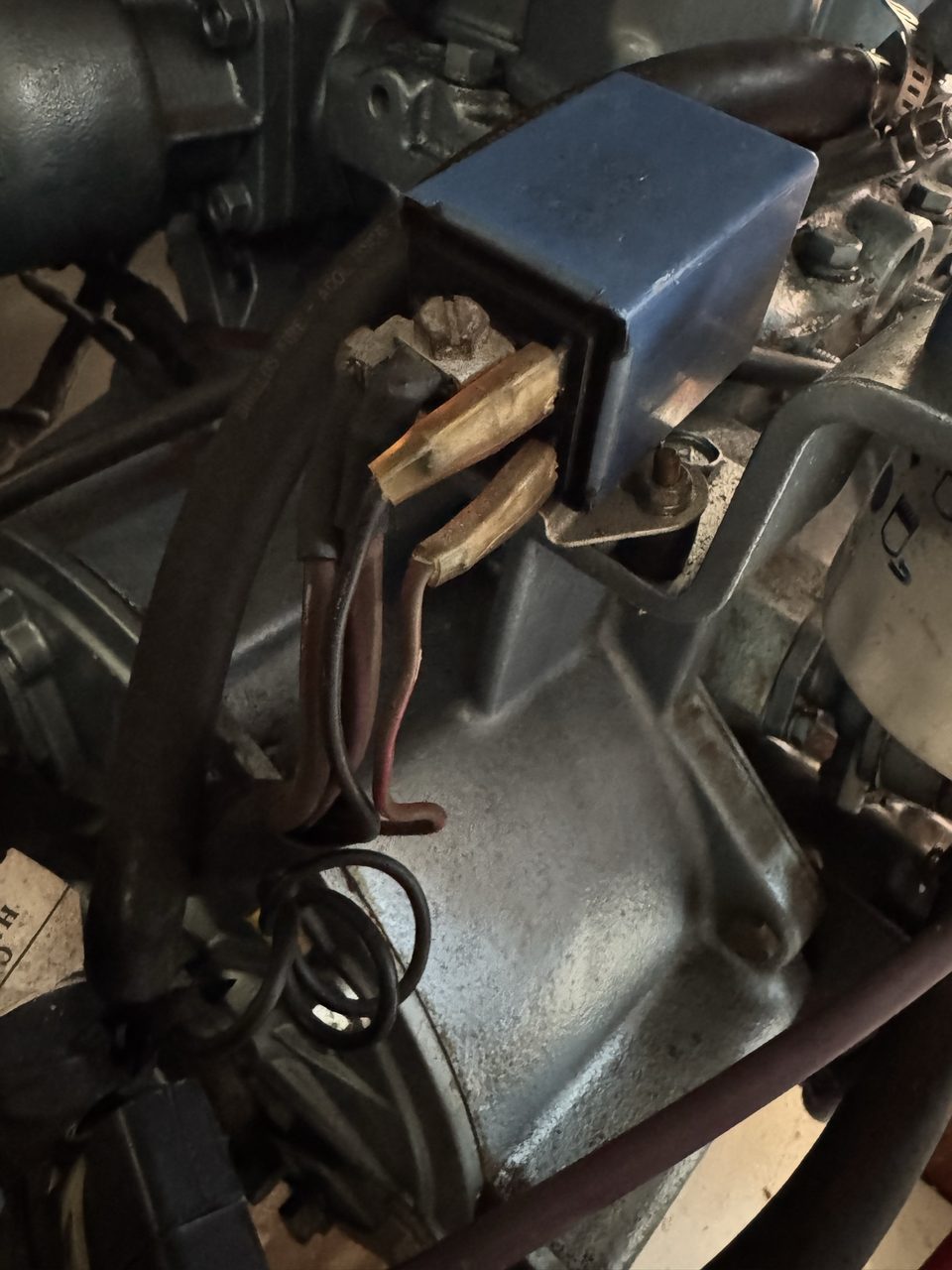 A blue plastic relay mounted on an engine bracket, with its cover lifted to show two heavily corroded brass screw terminals and thin brown wires