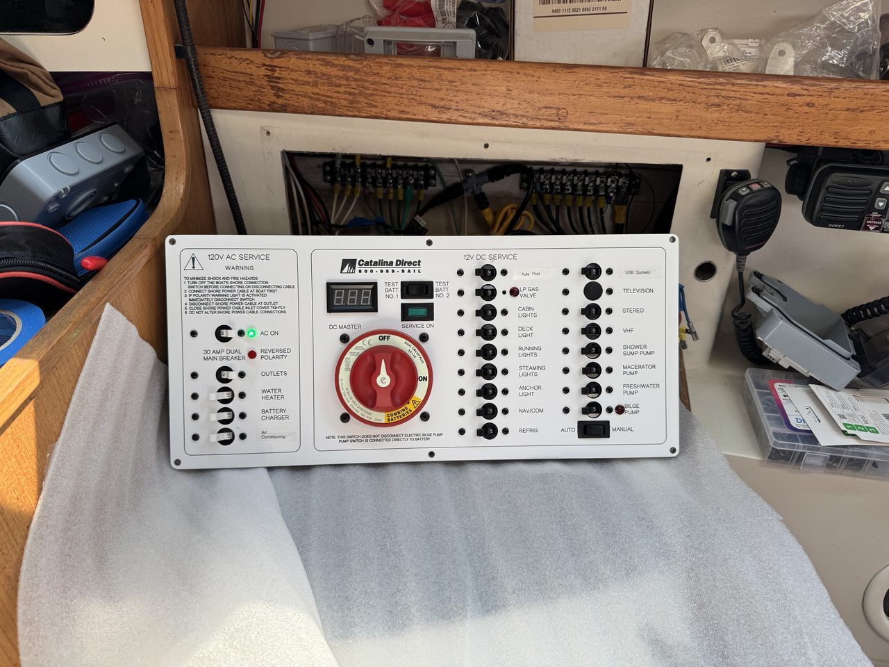 A new white marine electrical panel with rows of labeled breakers and a large red battery switch, lying on a foam pad in a boat cabin, with a small green indicator lamp lit