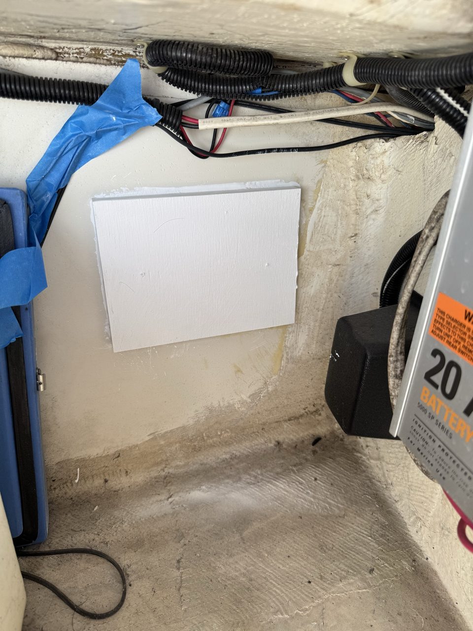 A white-painted rectangular plywood board bonded to the inside wall of a locker, with wiring in conduit above it and the label of a battery charger visible at the right