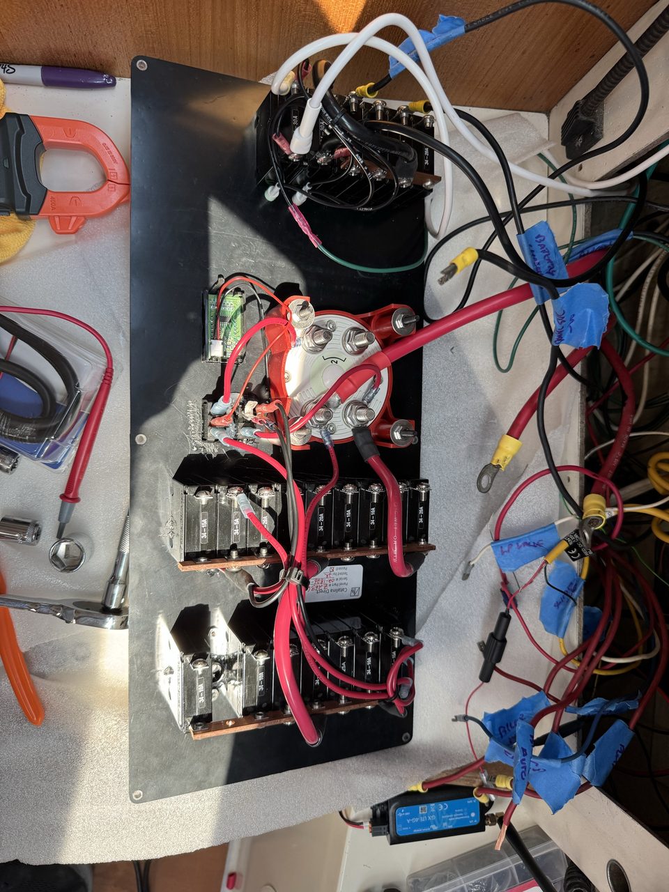 The reverse of a marine electrical panel showing rows of circuit breakers on copper busbars, a large battery switch, and a mass of red and yellow cables to one side, each tagged with blue masking tape