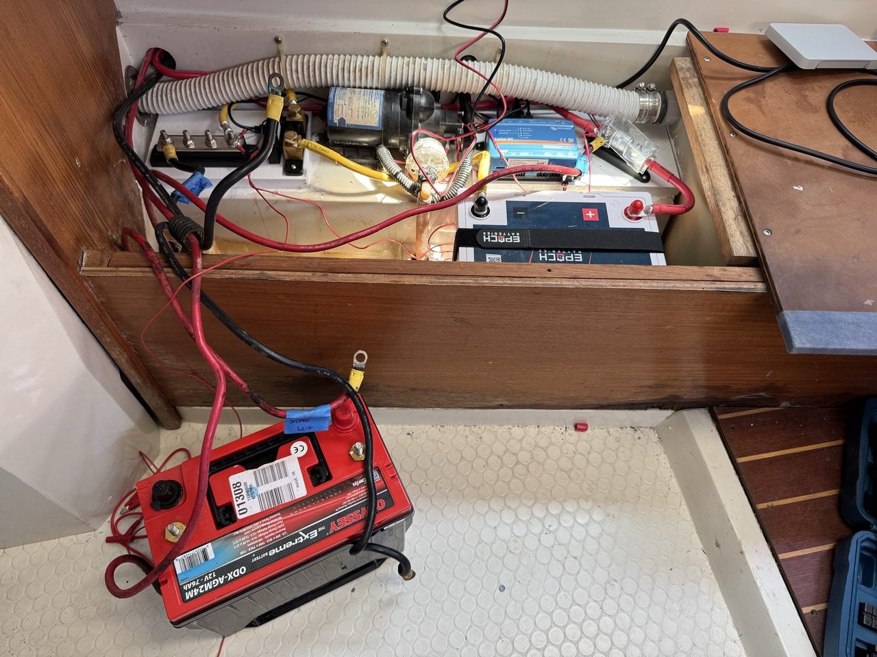 Looking into a boat battery compartment, with a lead-acid battery sitting out on a shelf, a lithium battery and cabling below, and one heavy black cable hanging unconnected with a yellow ring terminal on its end