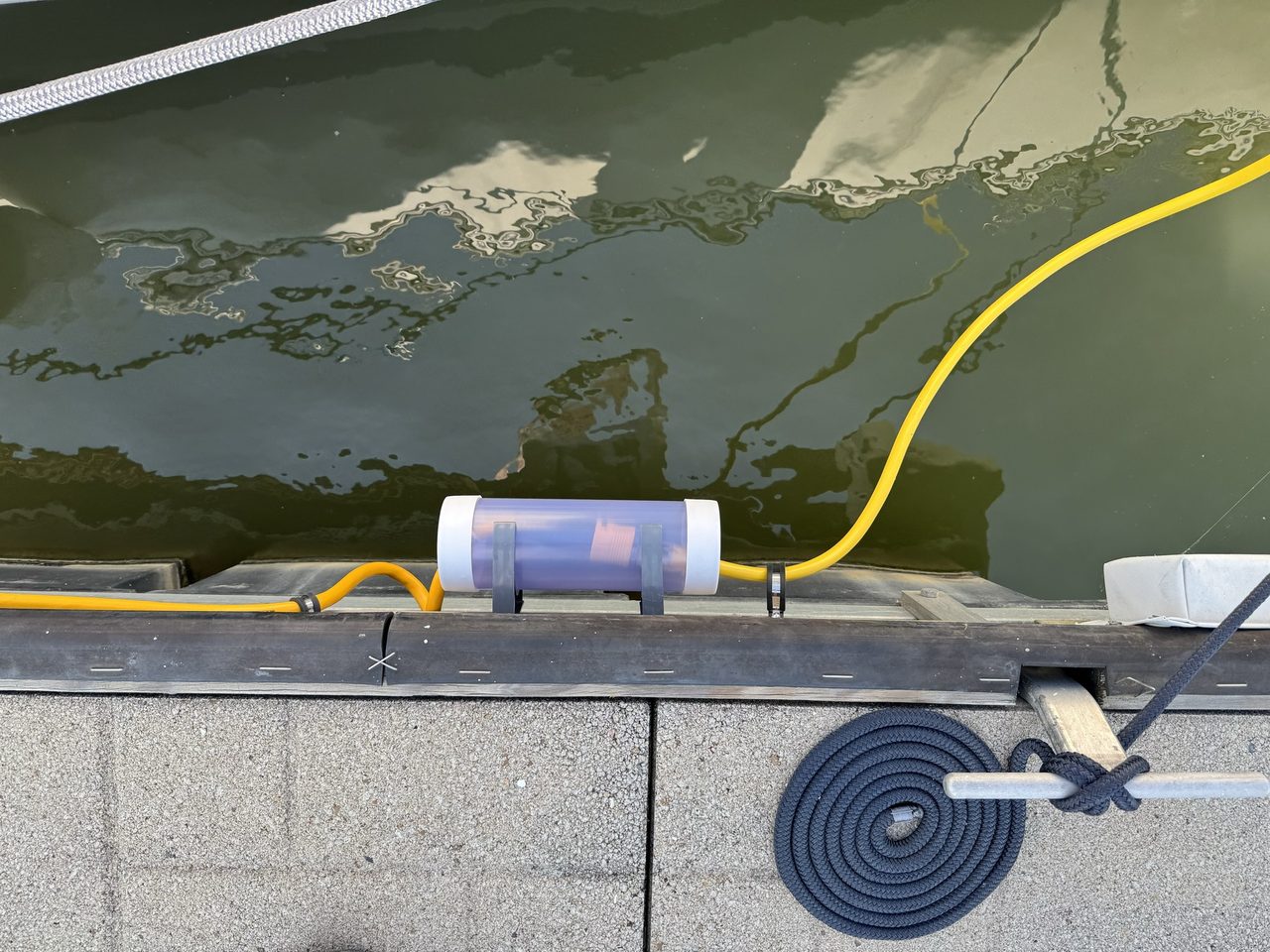 A length of clear conduit strapped to the side of the dock, the joint sheltered inside it, and the shore cord threaded through. The faint red glow inside the tube is the connector's own power-present light, which is why the tube is clear.